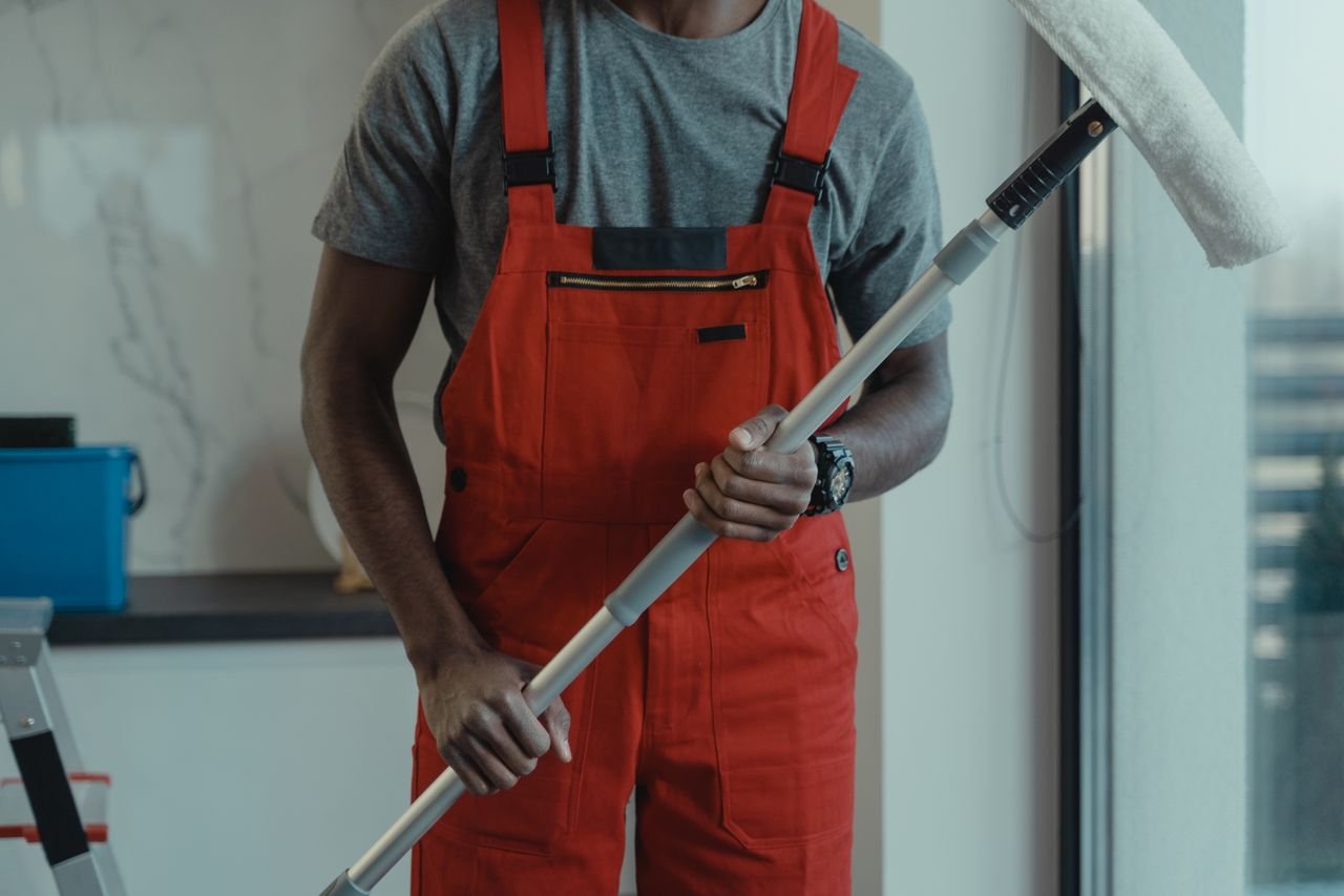 gallery-01 A professional cleaner wearing red overalls is holding a mop while cleaning indoors.